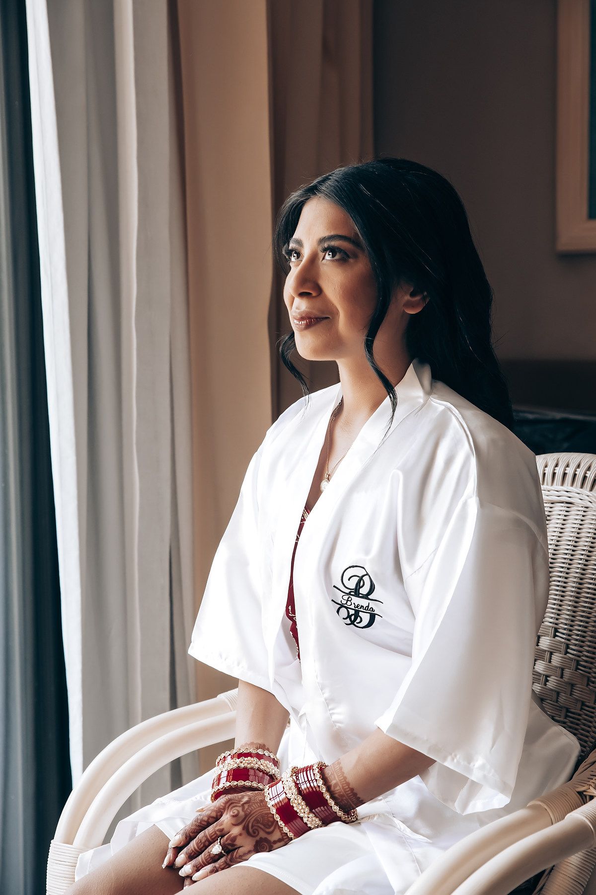 Bride in white monogrammed robe sitting by window with traditional henna and bangles.