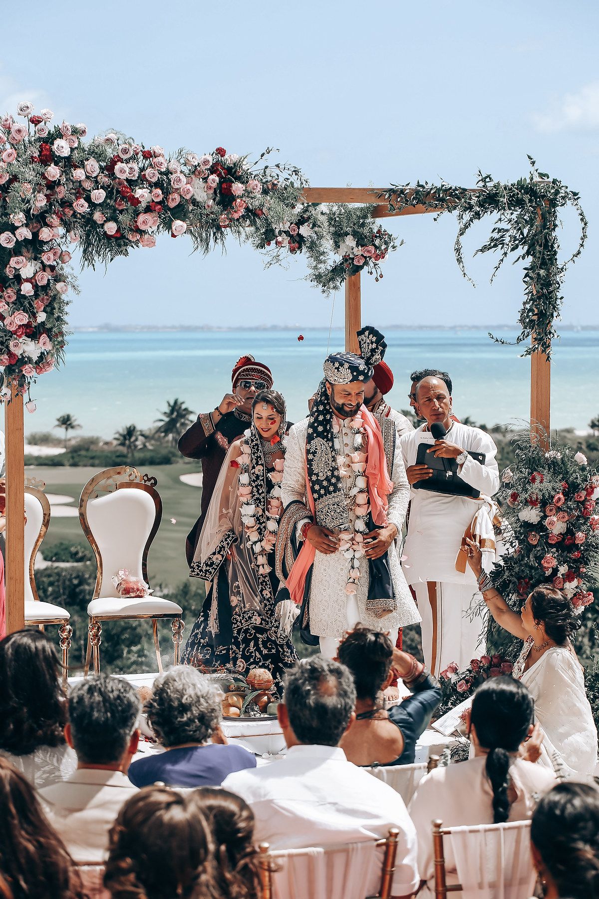 Wedding ceremony guests gather under floral decorations at a beachside venue.