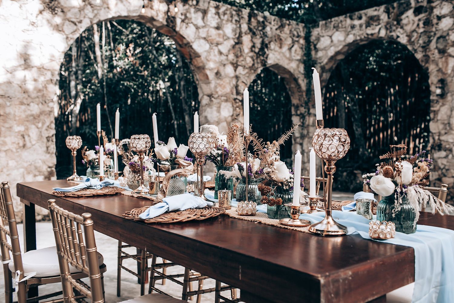 Elegant wedding reception tables decorated with white linens, floral centerpieces and candles in a rustic outdoor setting.