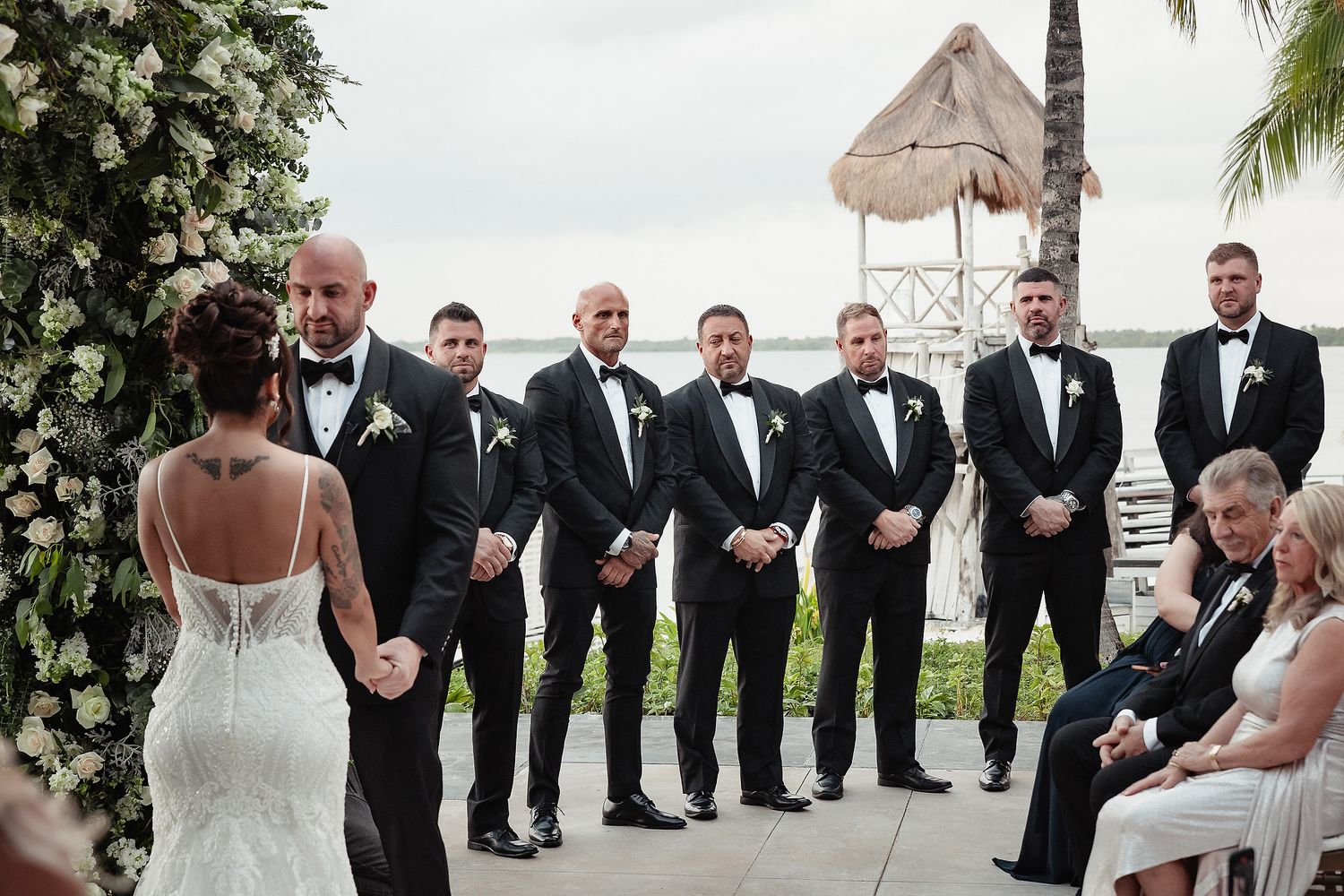 Wedding party in formal black tuxedos and white dress poses together on outdoor terrace with palm trees and greenery.