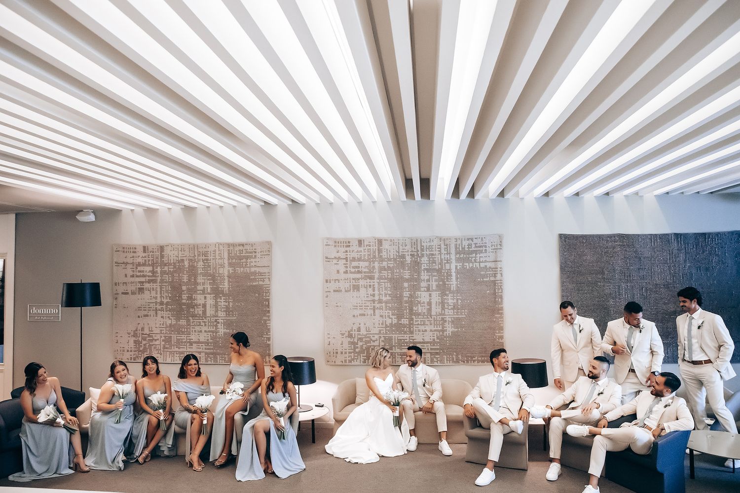 Wedding party members lined up in formal attire against a white architectural backdrop.