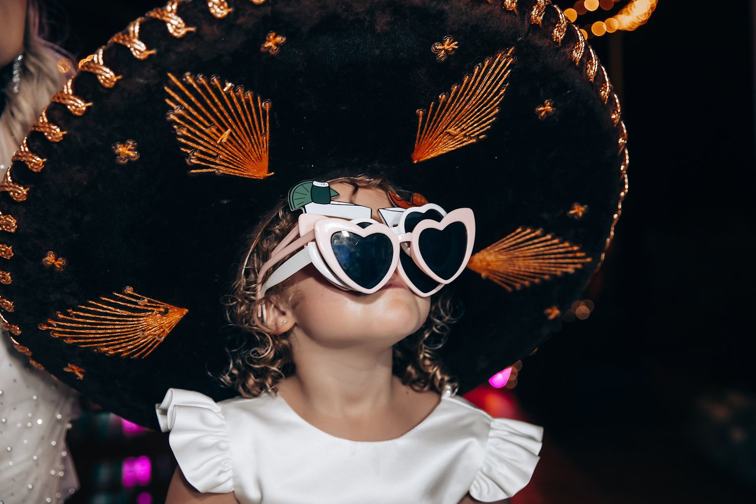 Partygoer wearing decorative Mexican sombrero and heart-shaped sunglasses at night celebration.