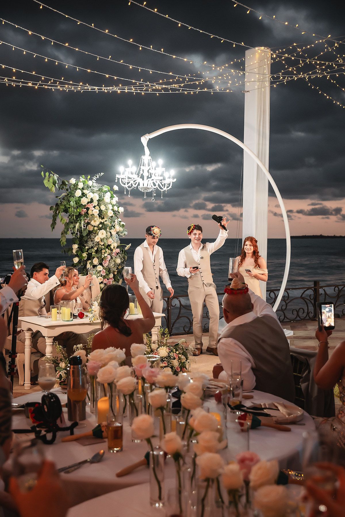 Evening beach wedding ceremony under string lights with floral arch and ocean view.