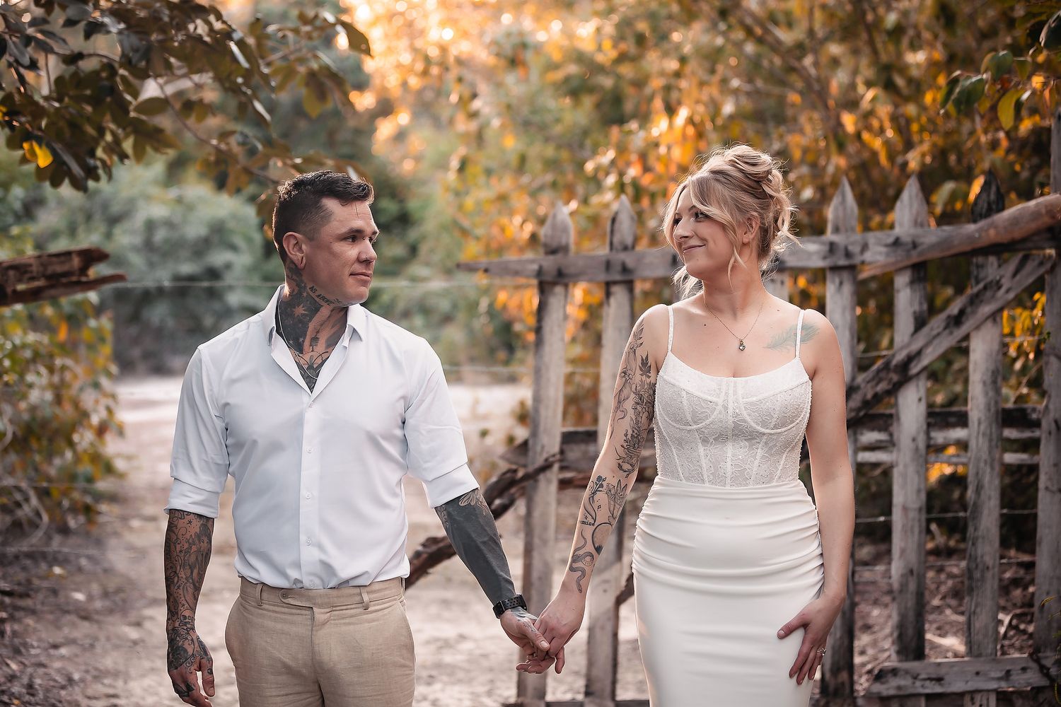 A couple walks hand in hand along an autumn path with rustic wooden fencing.