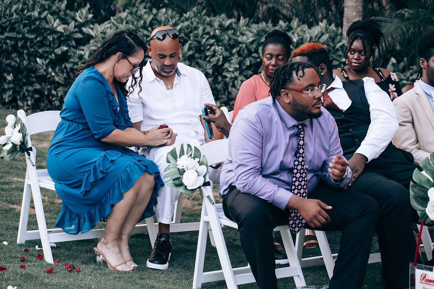 Guests seated in white chairs at an outdoor garden ceremony share joyful moments and laughter together.