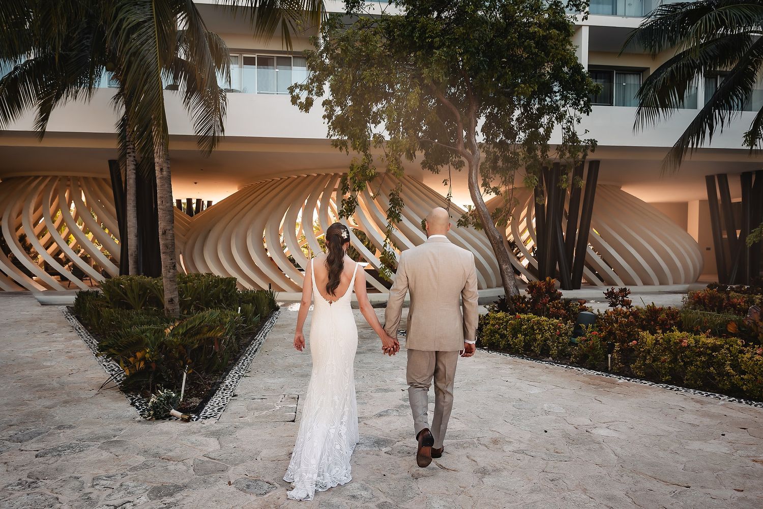 A couple walks hand in hand along a tropical resort path on their wedding day.