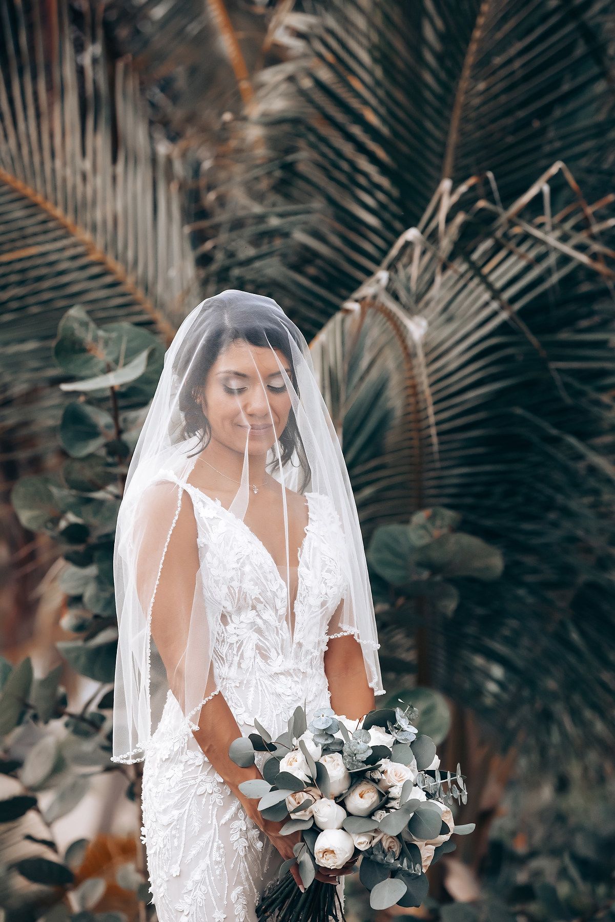 A white lace wedding dress and bouquet with eucalyptus leaves against a backdrop of palm fronds.