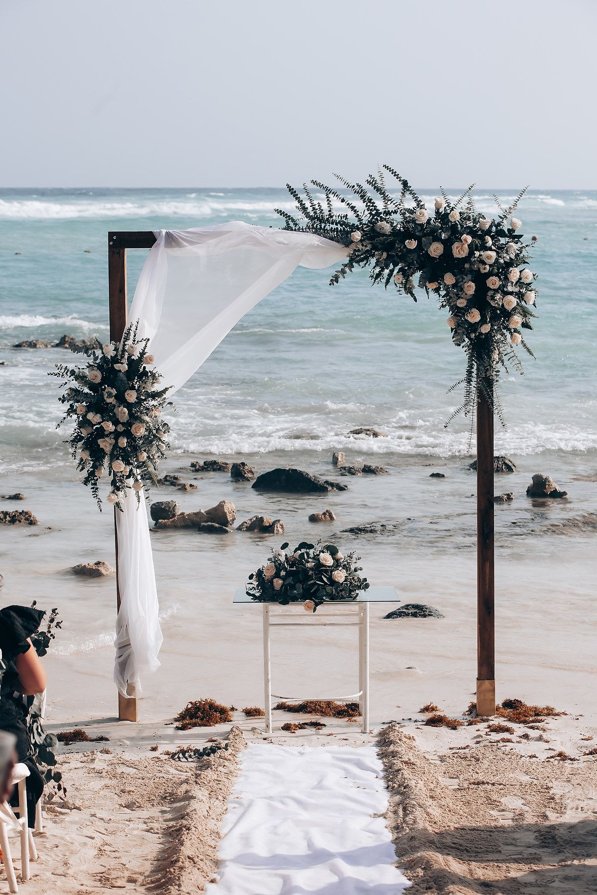 A beach wedding arch decorated with white fabric and dark florals stands against a dramatic ocean backdrop.