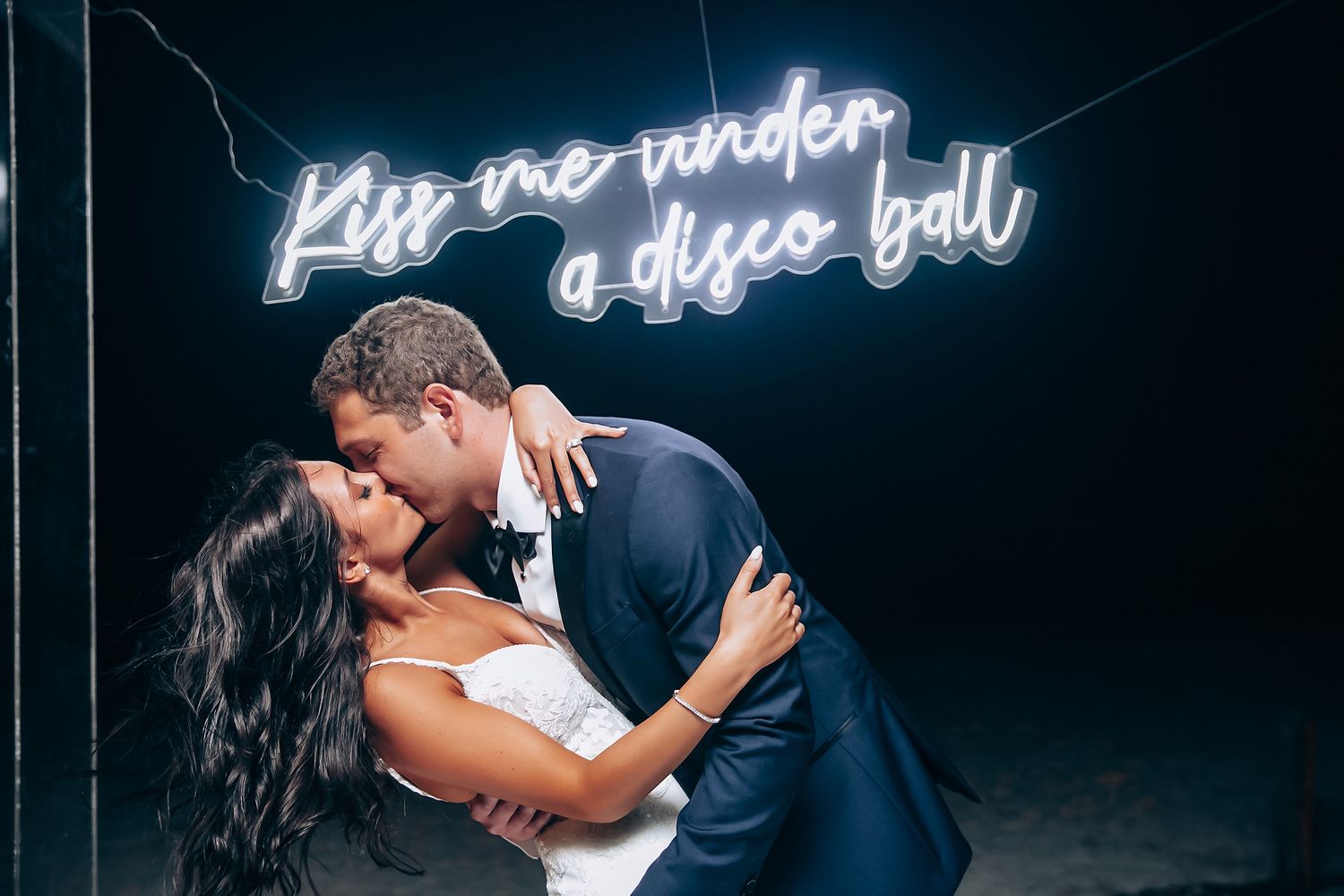 A romantic couple shares a passionate kiss at night under illuminated neon signage.