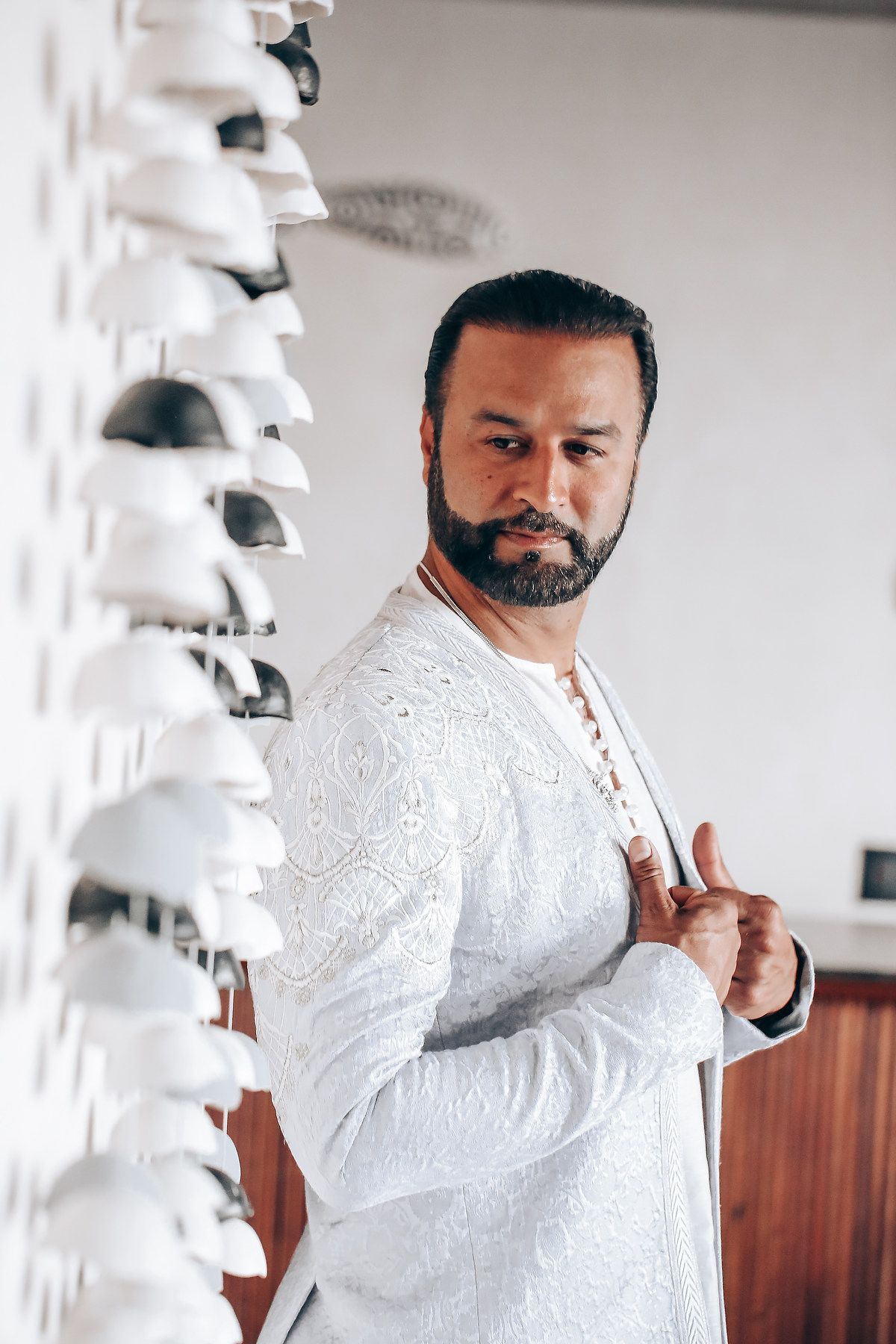 A bearded person wearing a white embroidered shirt stands next to a decorative white wall feature in an interior setting.