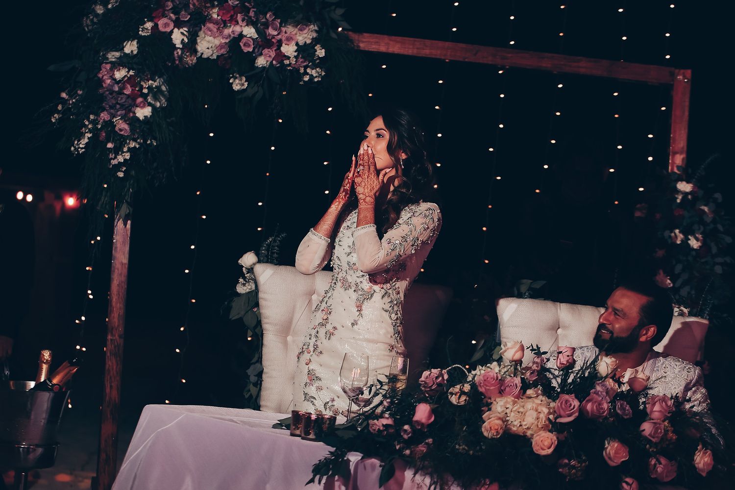 Elegant wedding ceremony setup with hanging lights, pink floral decorations, and white seating arrangements.