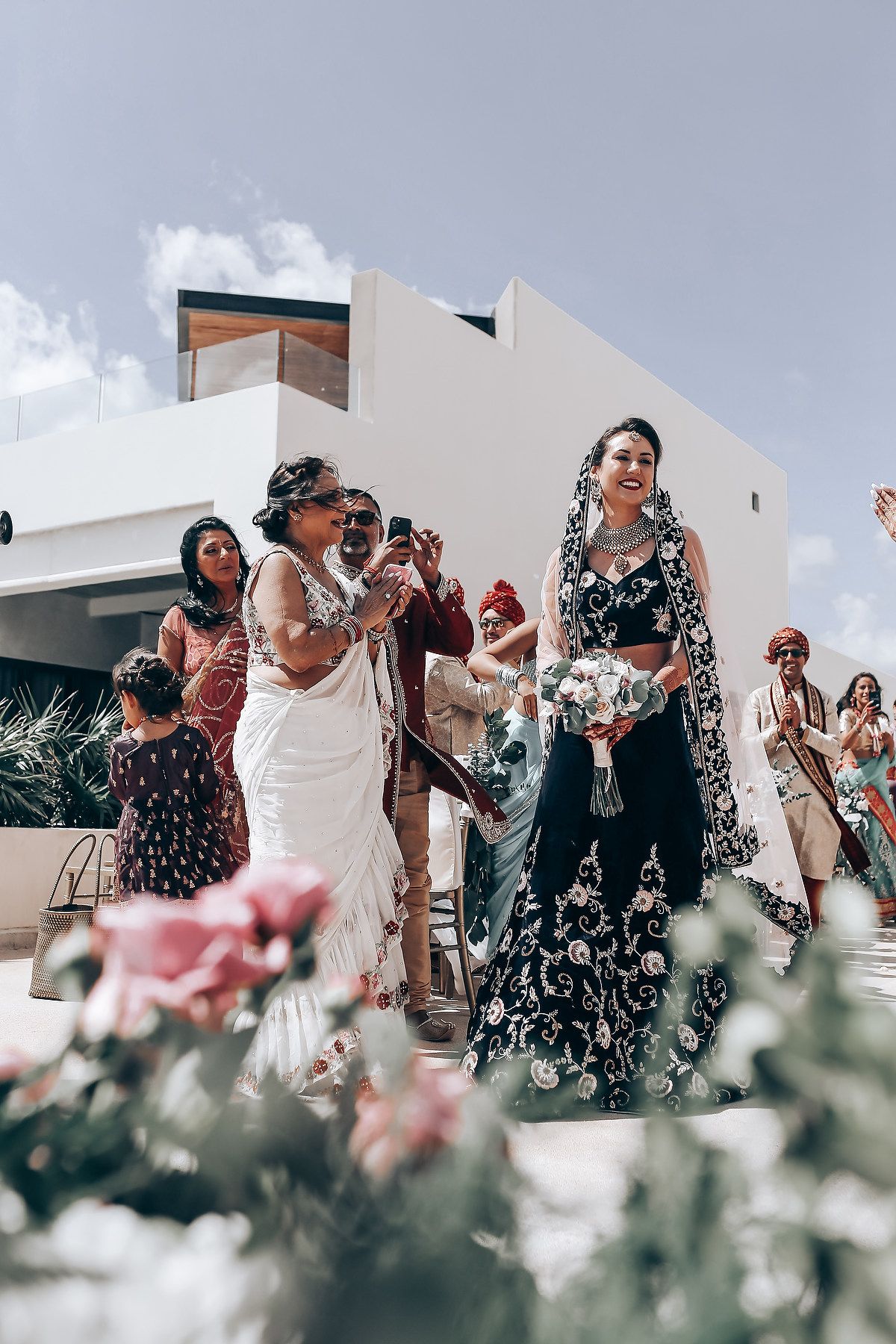 Wedding guests dancing and celebrating on an outdoor terrace with flowers in the foreground during a festive reception.