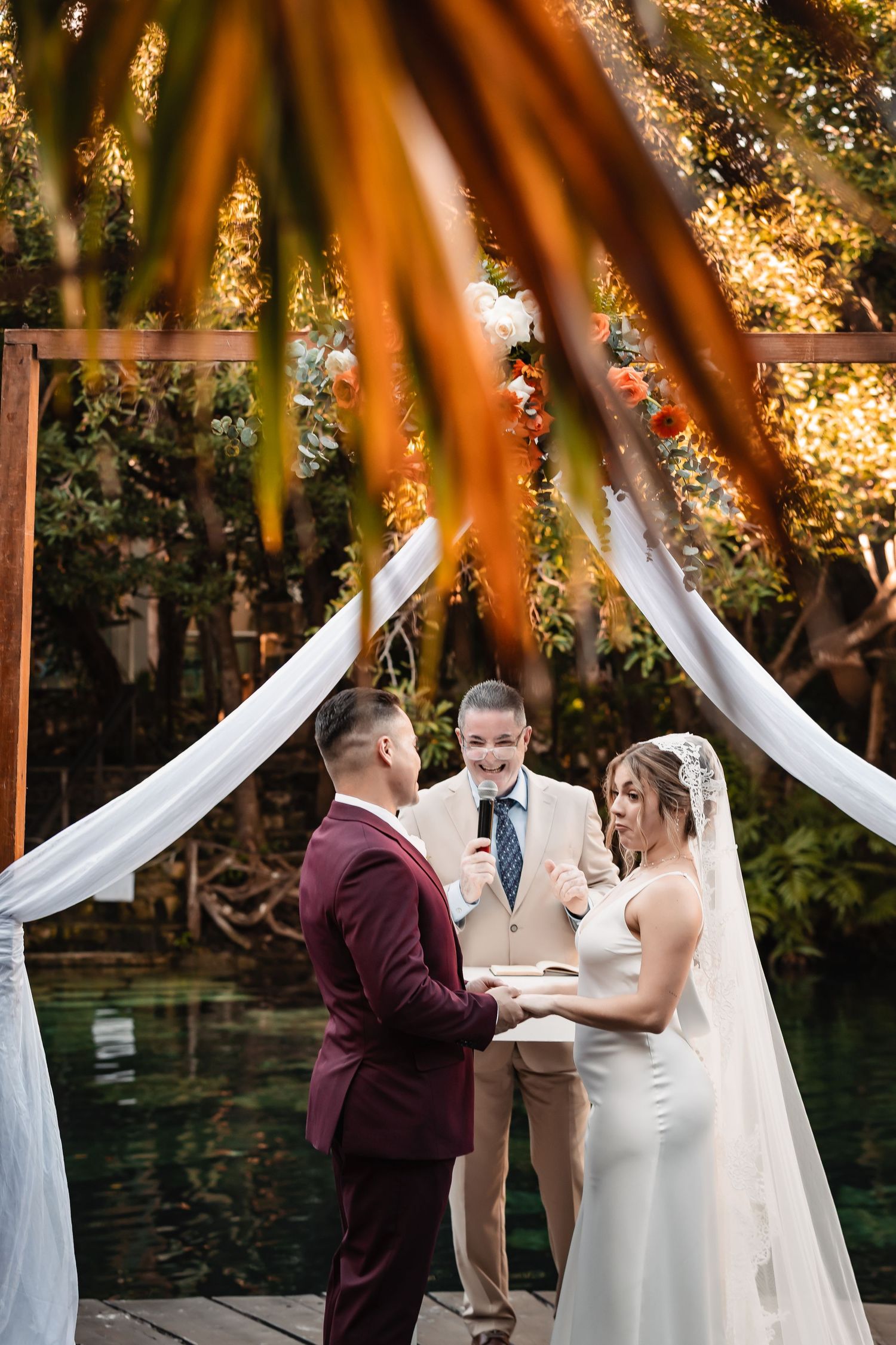 Wedding ceremony under floral arch with white draping and autumn foliage backdrop.