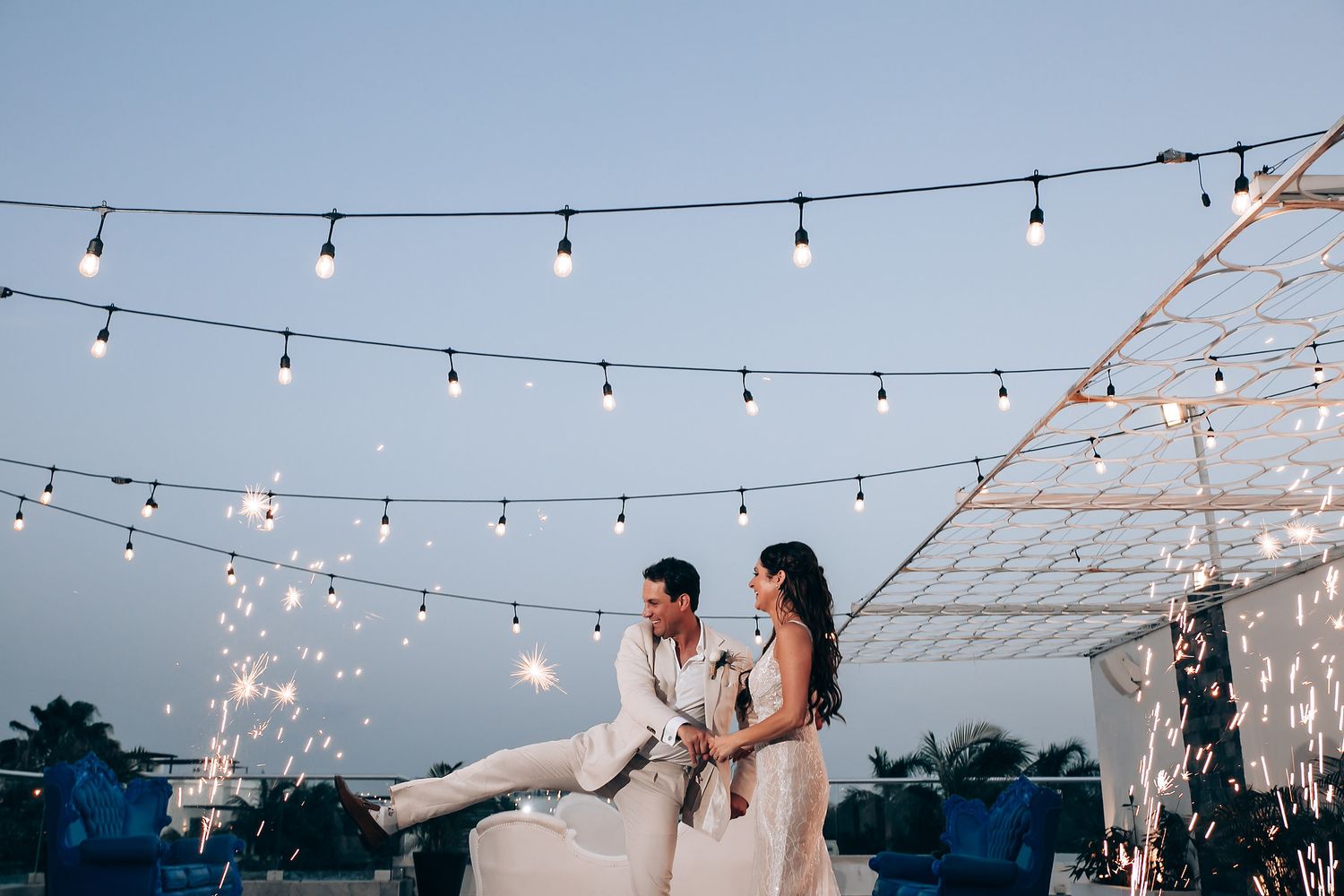 Couple sits on ledge during twilight celebration with string lights and sparkler effects.