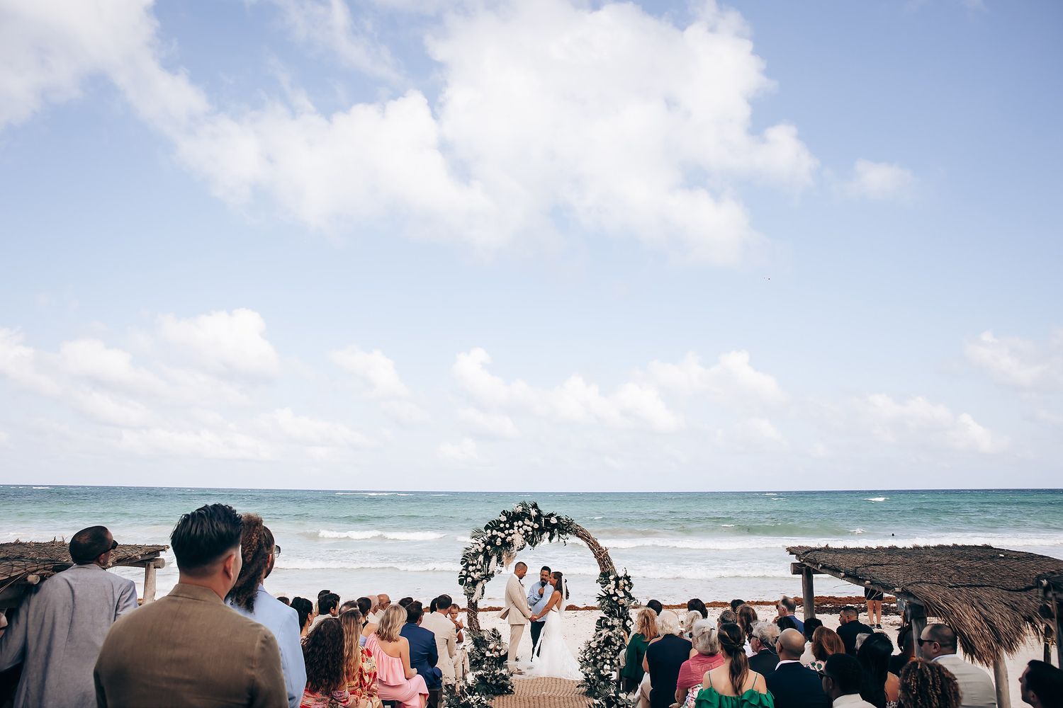 Oceanfront wedding ceremony with guests viewing under partly cloudy skies.