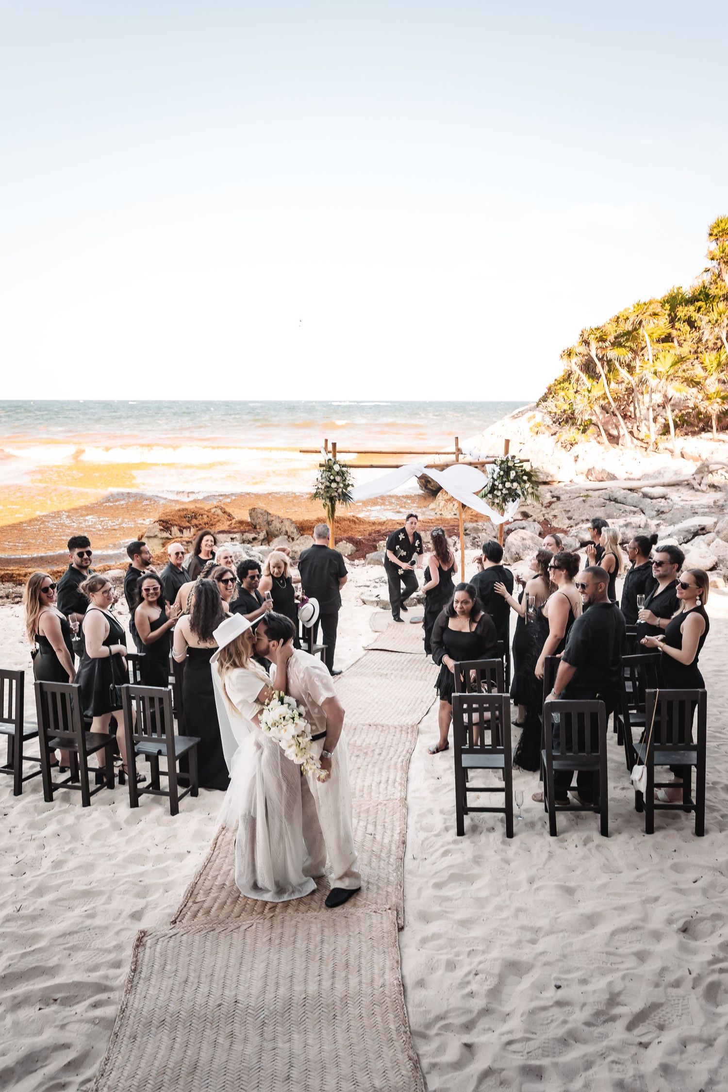 Beach wedding ceremony with guests seated in black chairs on white sand.