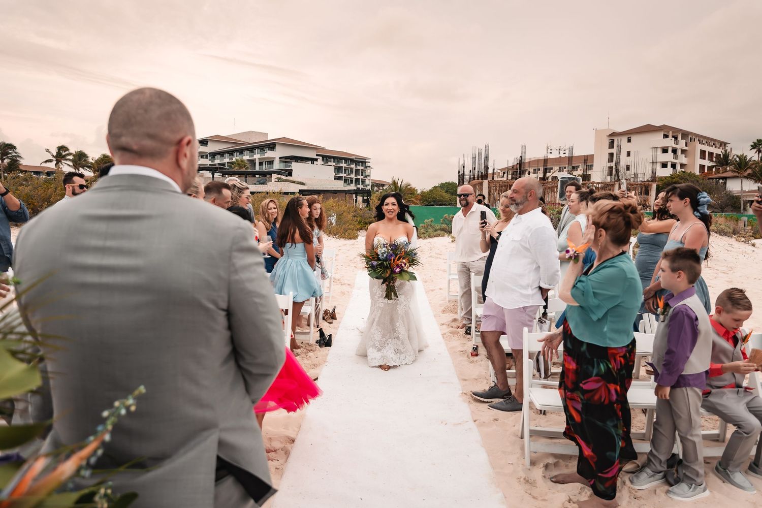 A romantic beach wedding ceremony with guests watching under a cloudy sky.