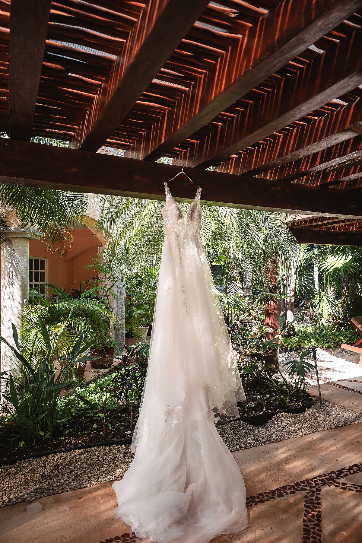 White wedding dress hanging under wooden pergola surrounded by tropical garden landscaping.