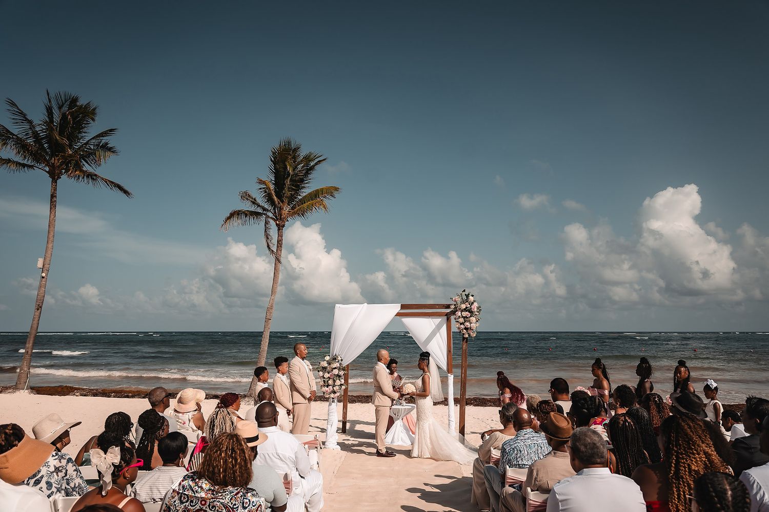 Panoramic shot of beachside wedding ceremony with palm trees and ocean backdrop.