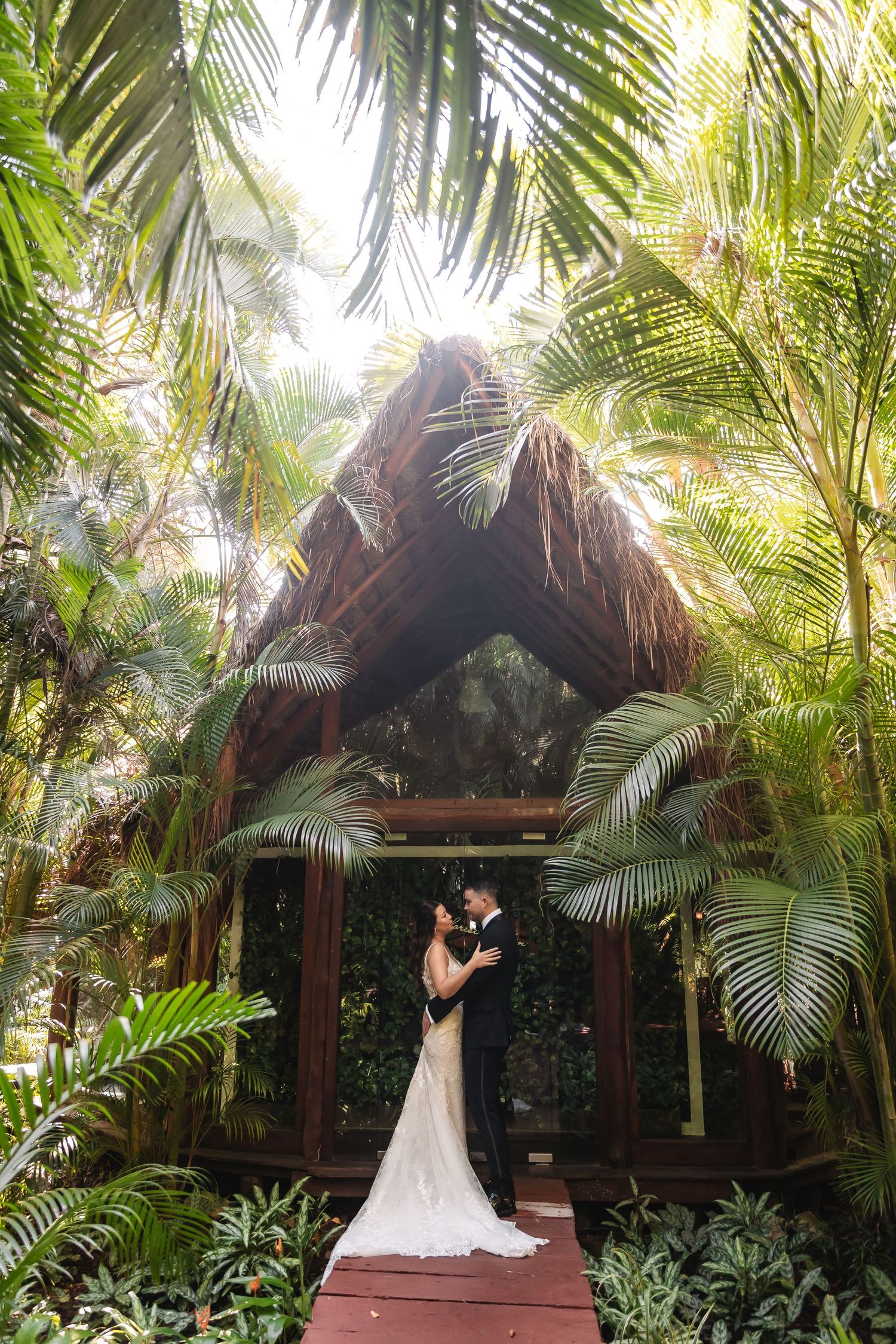Wedding couple poses under a thatched roof pavilion surrounded by tropical palm trees.