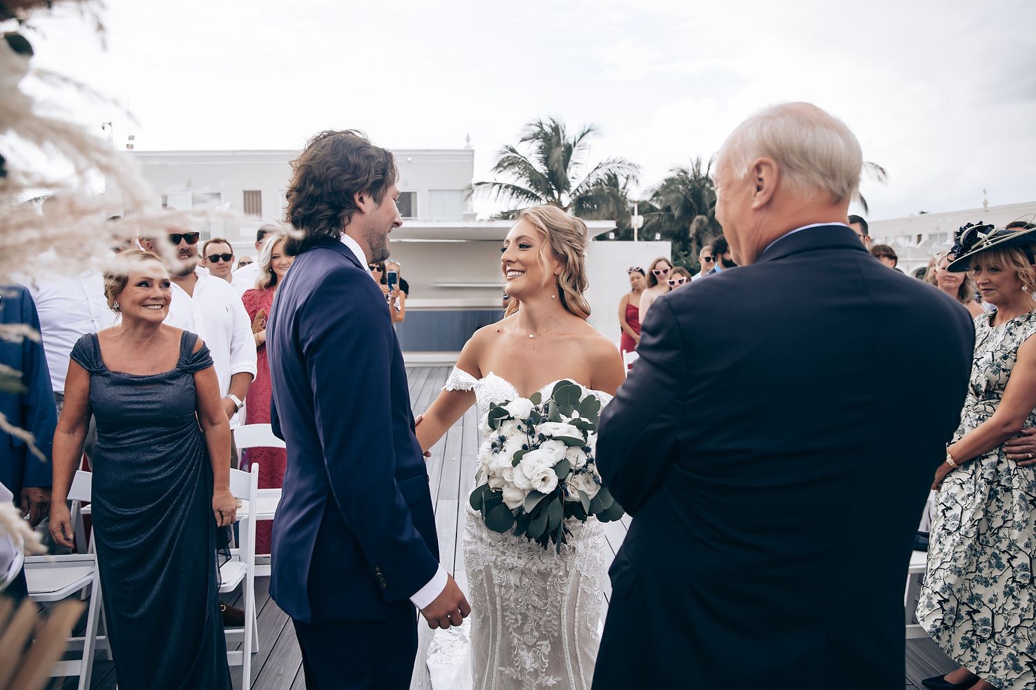 Wedding guests seated in white chairs at an outdoor ceremony by the ocean.