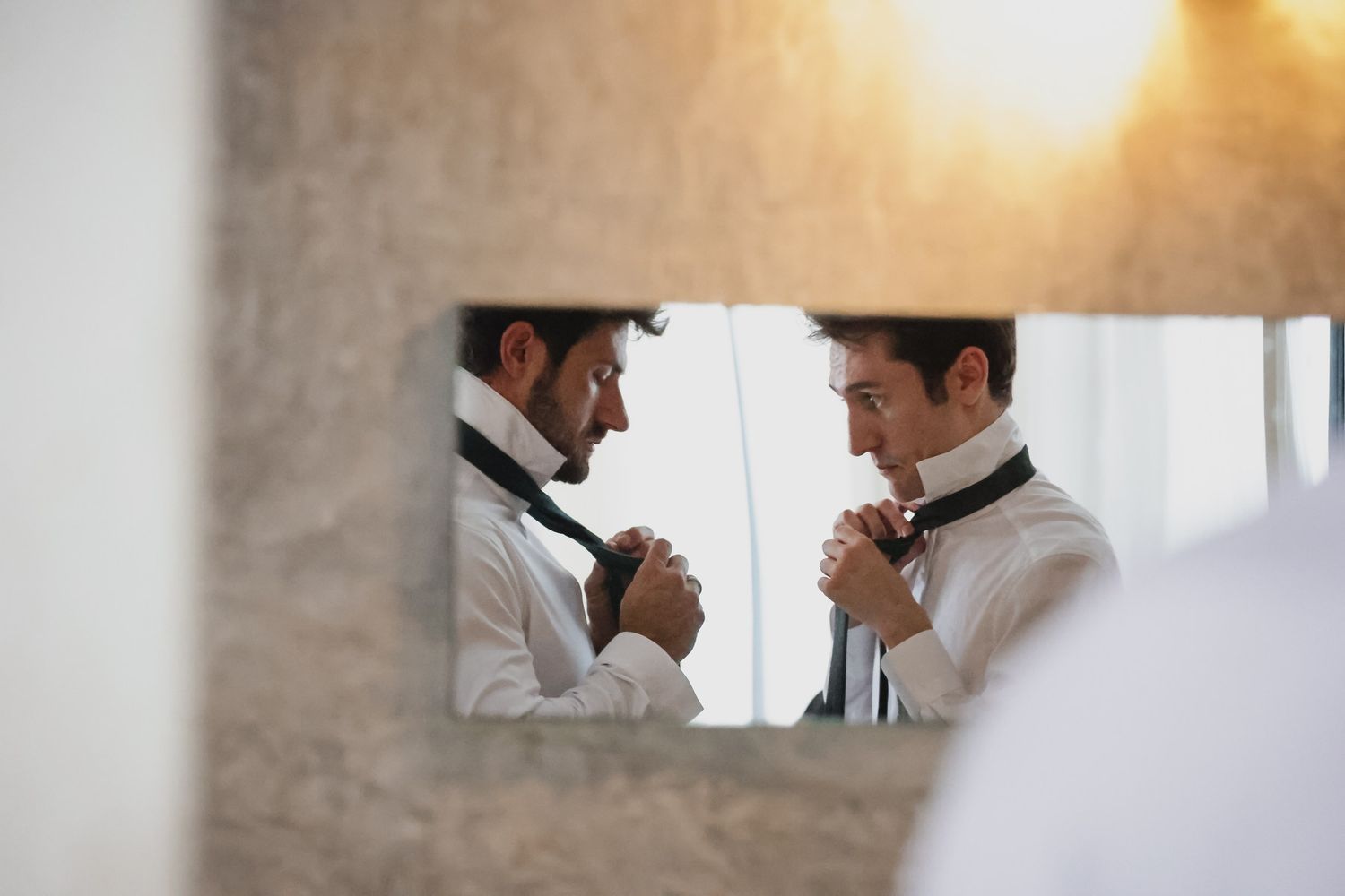 Man adjusting black tie in mirror reflection, wearing white dress shirt, warm golden backlight, wedding preparation scene.
