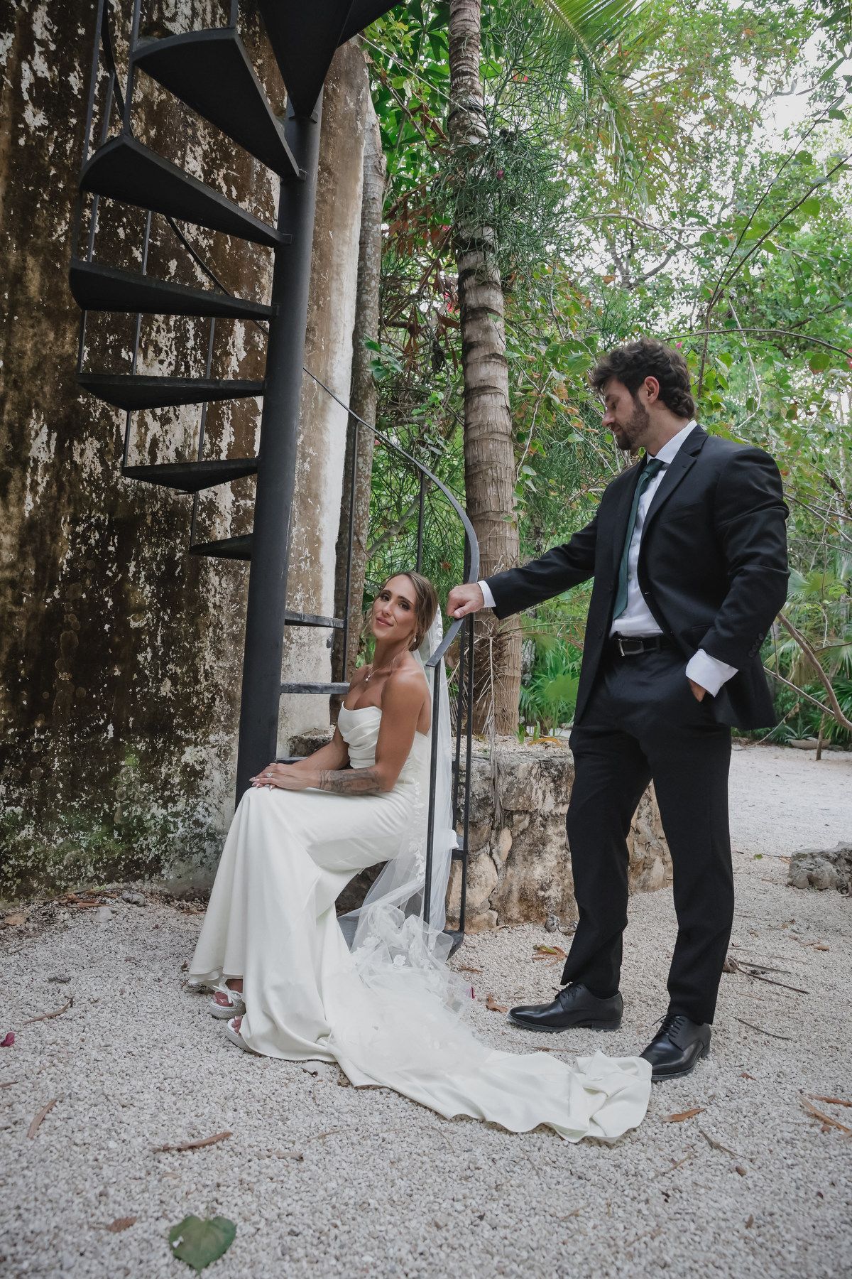 Bride in white gown sits on spiral staircase as groom in black suit stands beside her in lush outdoor setting.