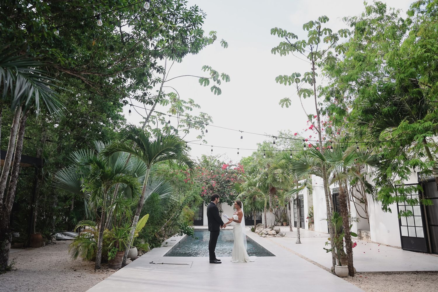 A bride and groom share a romantic moment in a lush tropical courtyard with string lights and a reflecting pool.