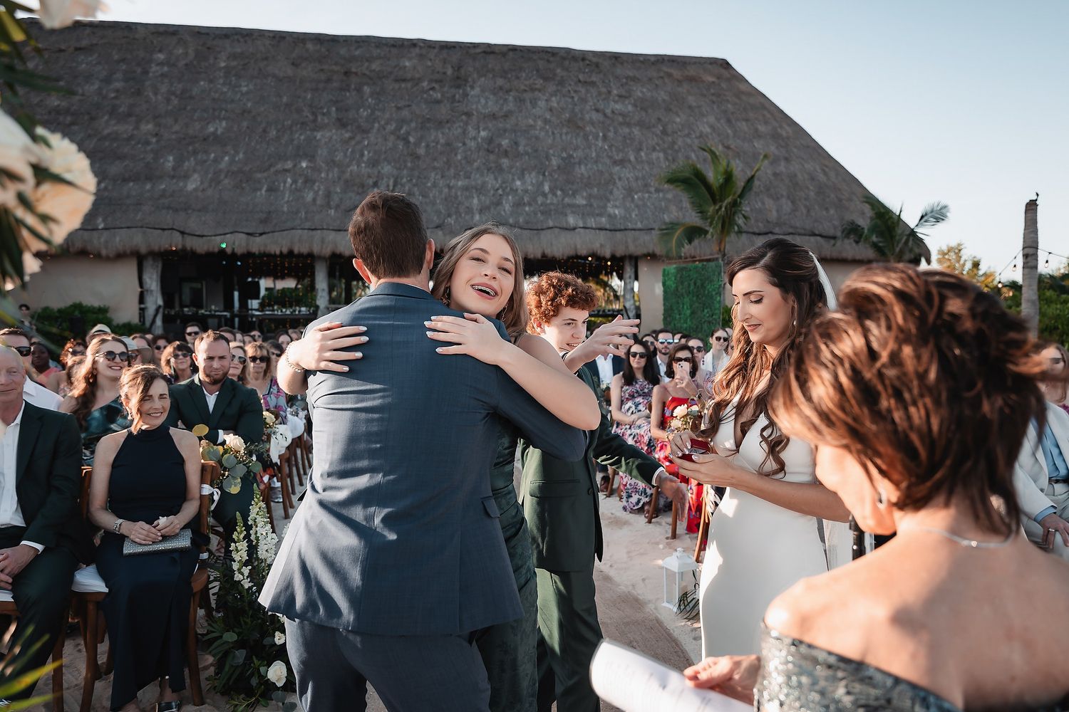 Bride and groom share a joyful hug during outdoor wedding ceremony at tropical venue with thatched roof.