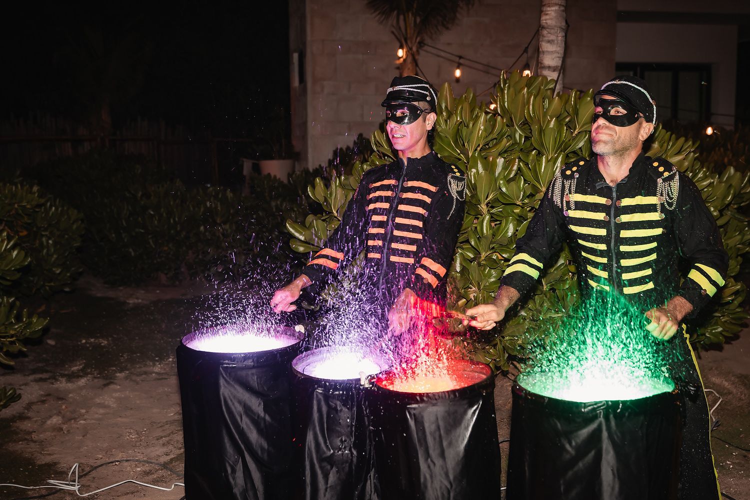 Two masked performers in marching band jackets create colorful smoke effects from illuminated cauldrons at a nighttime event.