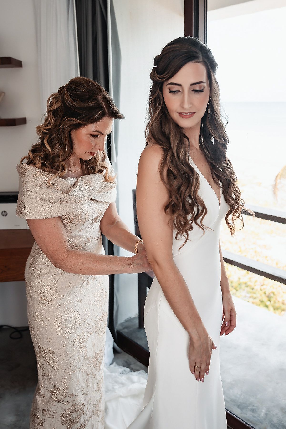 A bride in a white gown gets help from a woman in lace dress, preparing for her wedding near a scenic window.