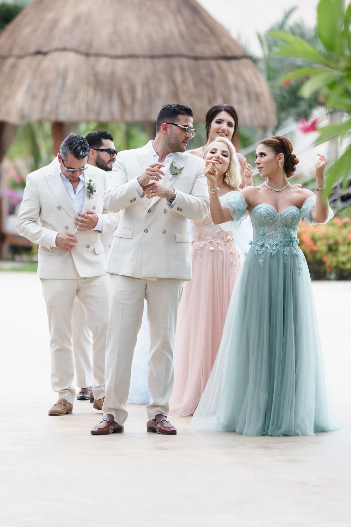 Wedding party in elegant attire, groomsmen in cream suits and bridesmaids in pink and blue gowns at tropical venue.