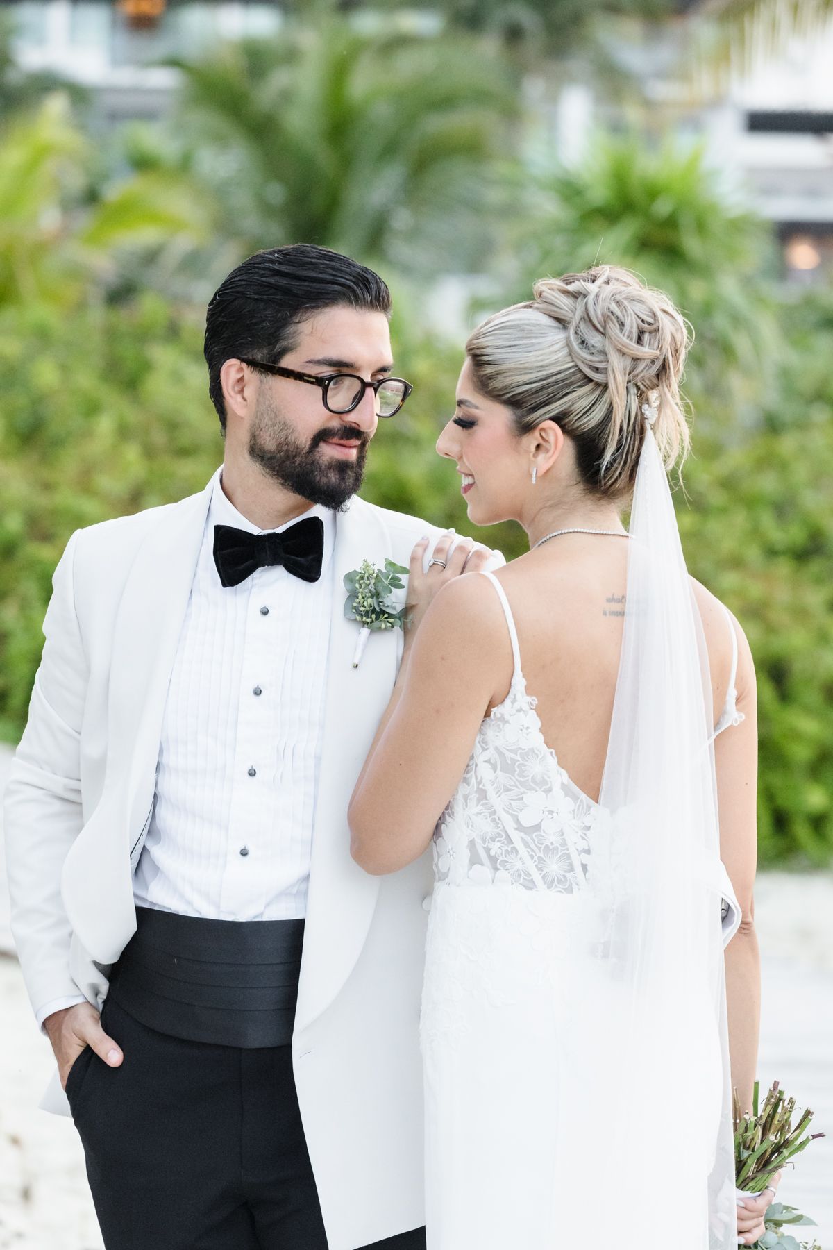 Groom in white tuxedo and bride in backless gown share a romantic moment outdoors.