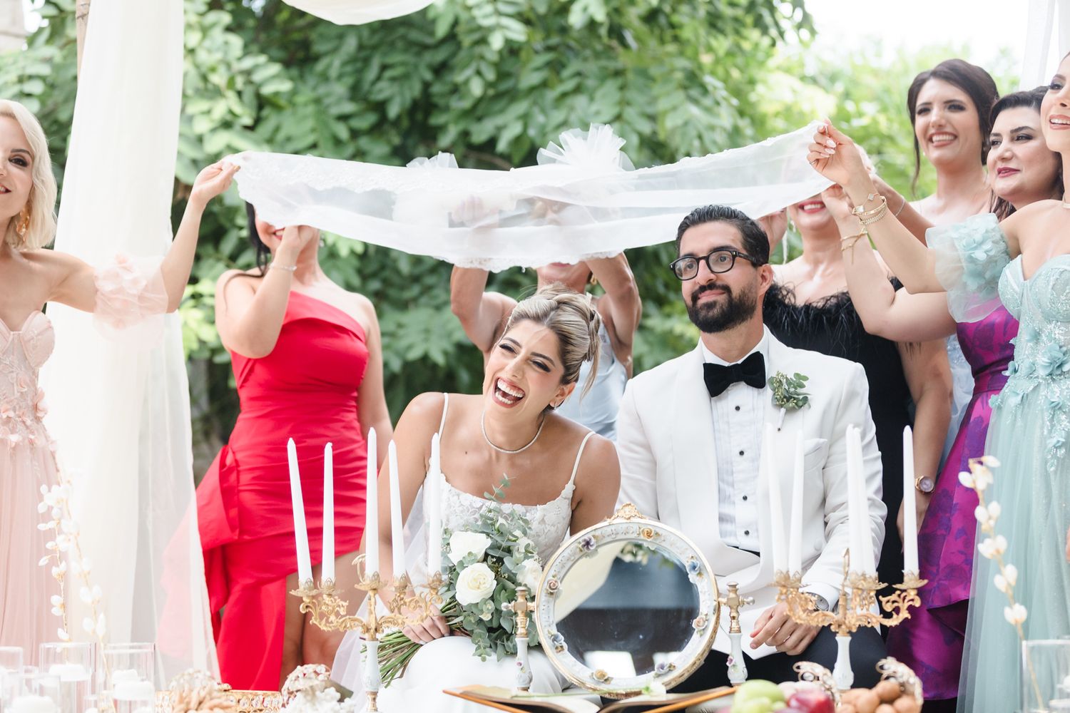 Joyful bride and groom seated at wedding table while bridesmaids in colorful dresses hold white veil overhead outdoors.