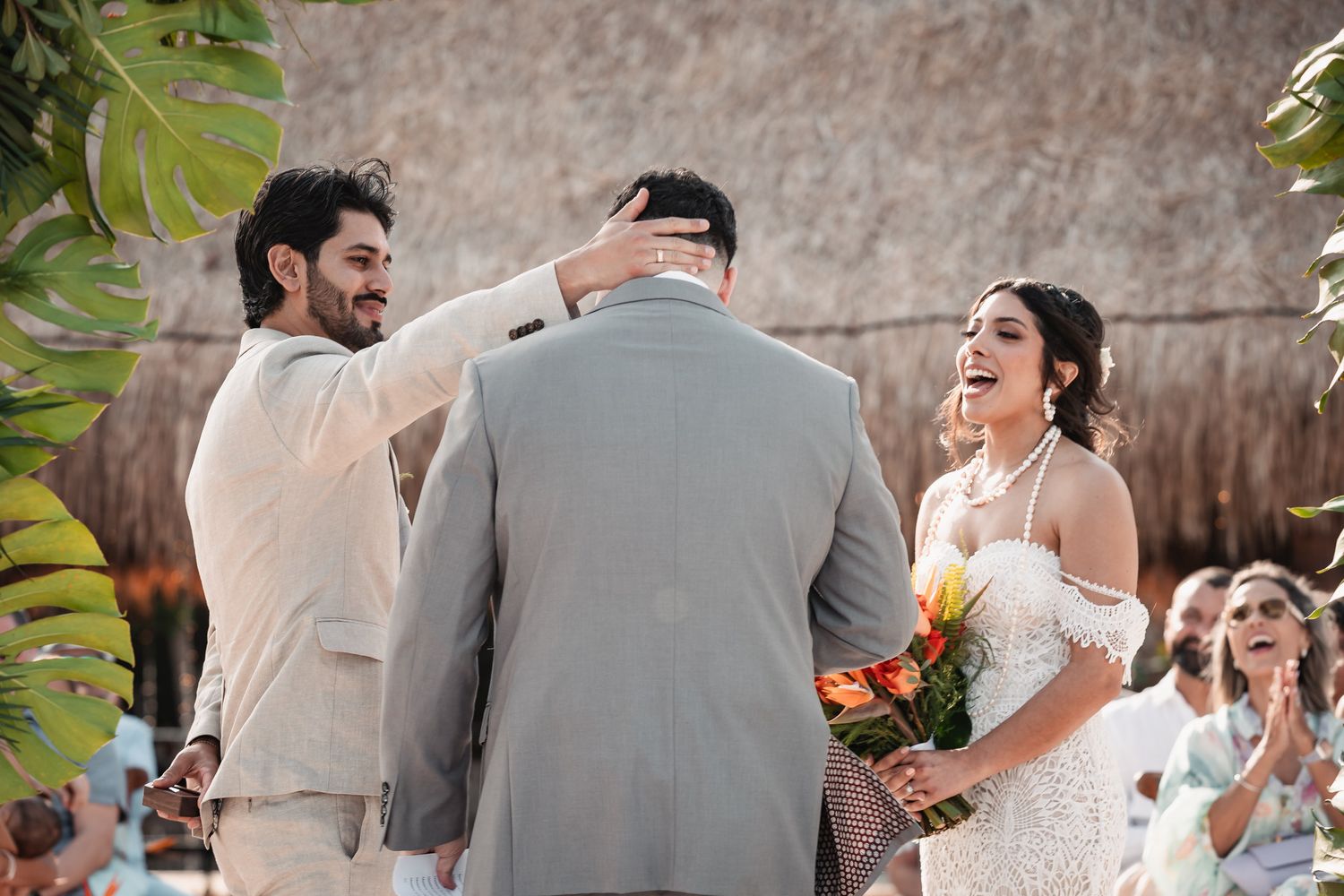 Groom receives a blessing from officiant as laughing bride holds tropical bouquet at beach wedding.