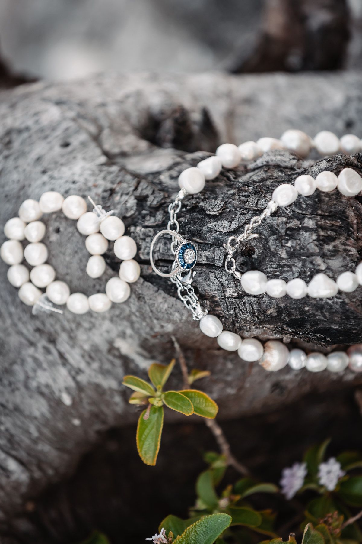 Elegant pearl necklace, bracelet, and silver ring with blue gemstone displayed on weathered driftwood outdoors.