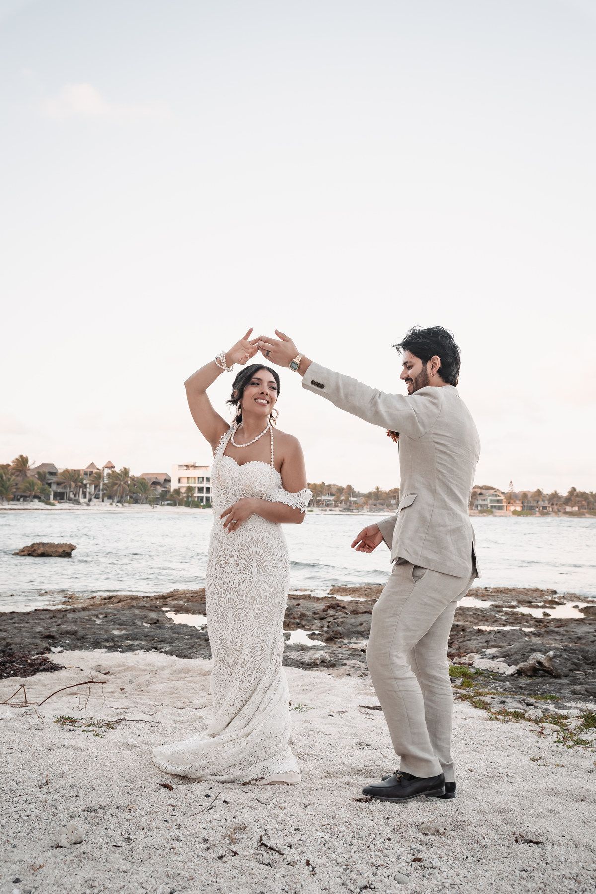 Bride and groom dancing joyfully on rocky beach at twilight by the sea.