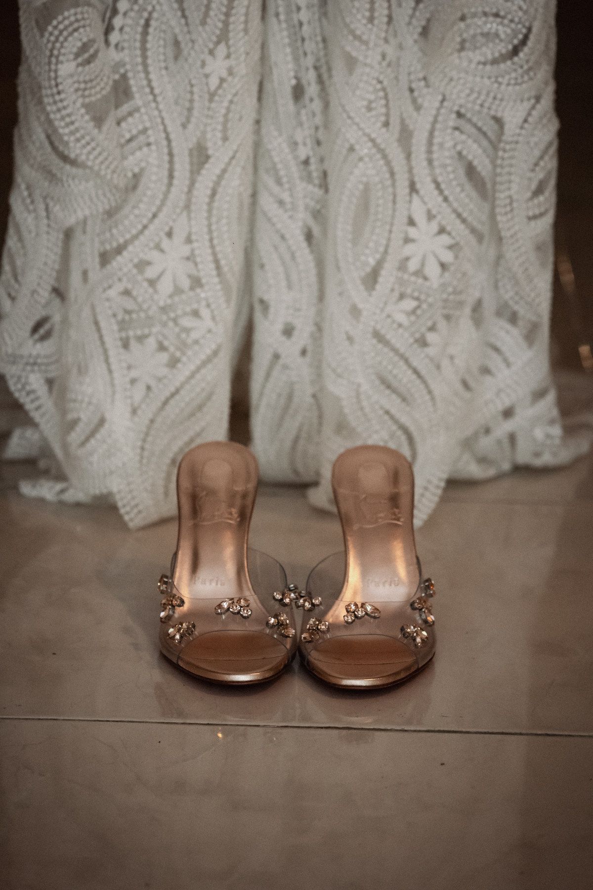 Elegant rose gold bridal heels with jewel embellishments displayed in front of a white lace wedding dress.