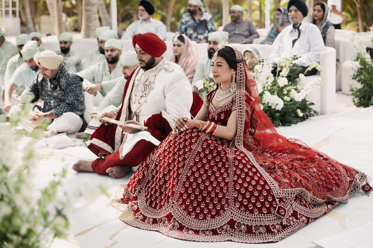 Sikh bride and groom in traditional red and white attire during beach wedding ceremony.
