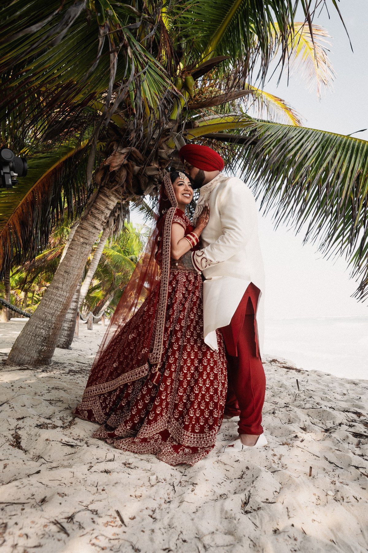 Indian bride and groom in traditional red and white wedding attire embracing under tropical palm tree.