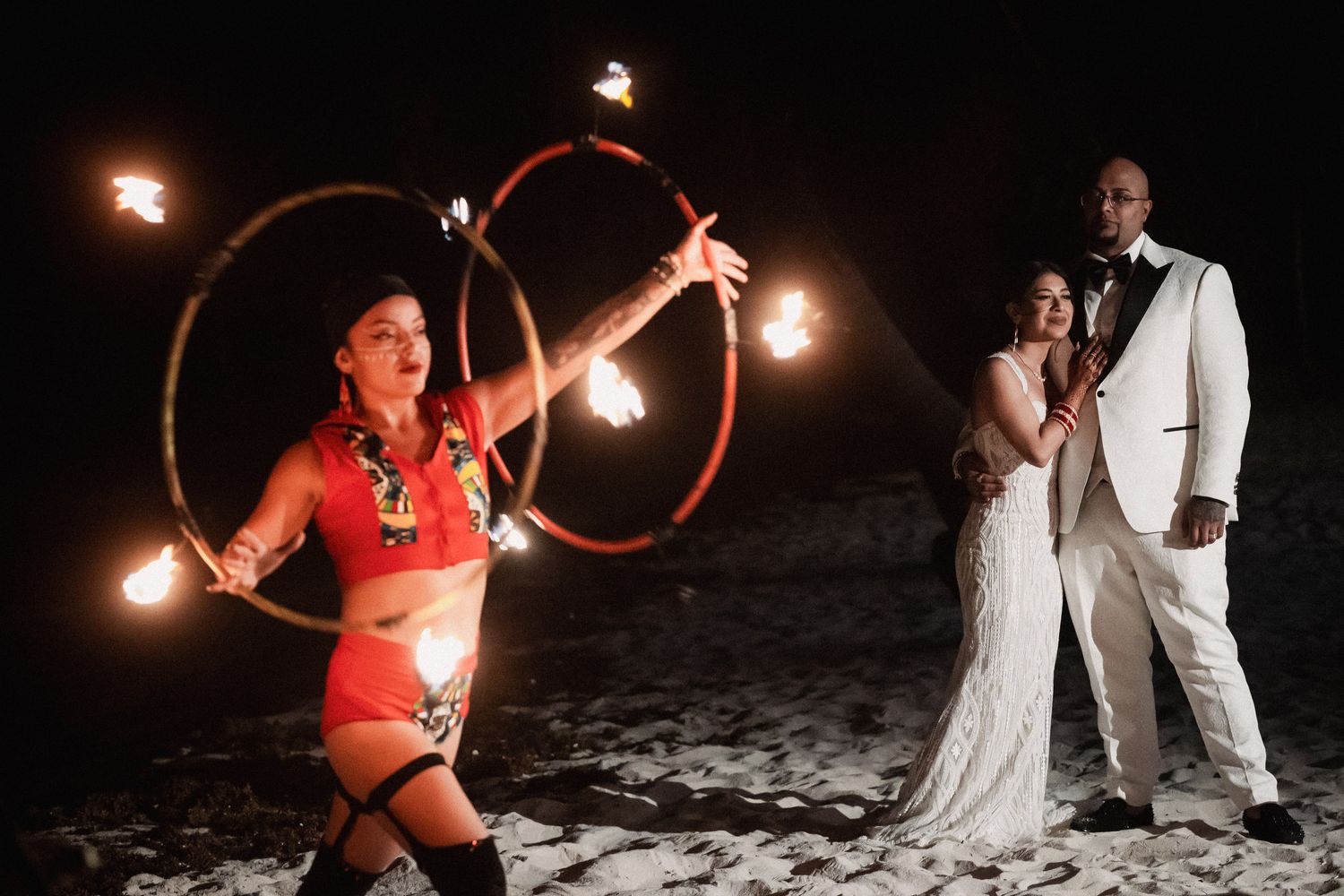 Bride and groom watch fire dancer performing with flaming hoops on beach at nighttime wedding celebration.