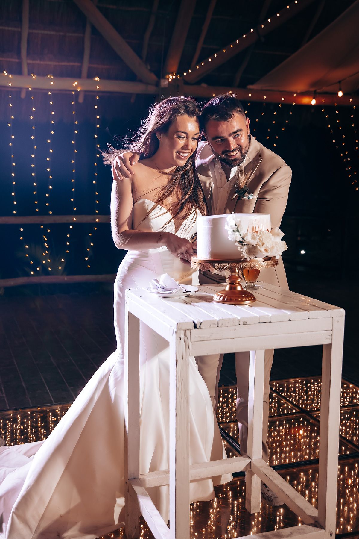 Couple shares intimate moment at white desk with fairy lights creating romantic ambiance in background.
