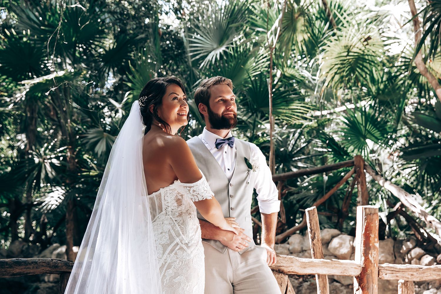 A romantic wedding couple shares a joyful moment under palm trees in a tropical outdoor setting.