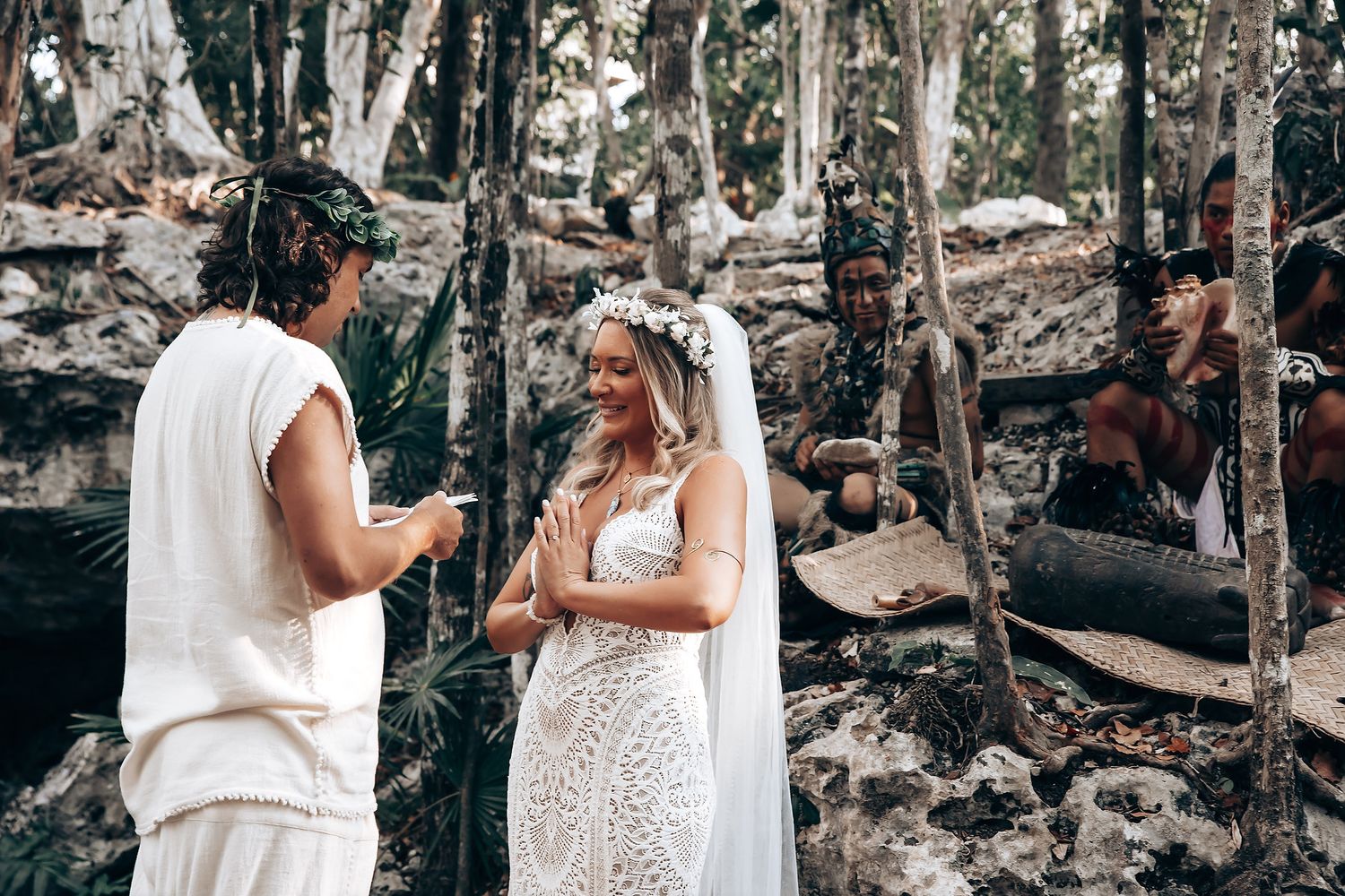 A couple shares an intimate moment in a wooded outdoor setting wearing white attire.
