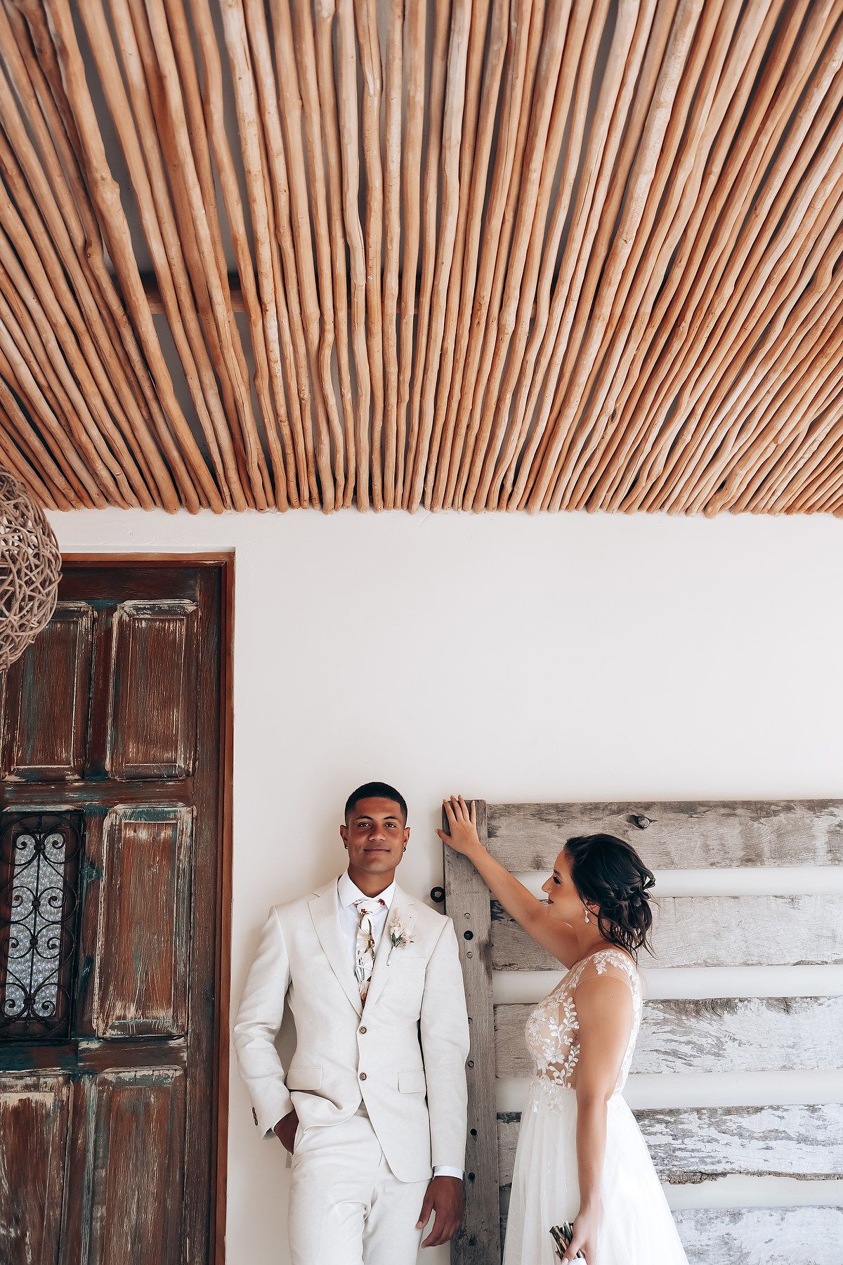 Wedding couple under rustic bamboo ceiling with textured white walls and vintage wooden doors.