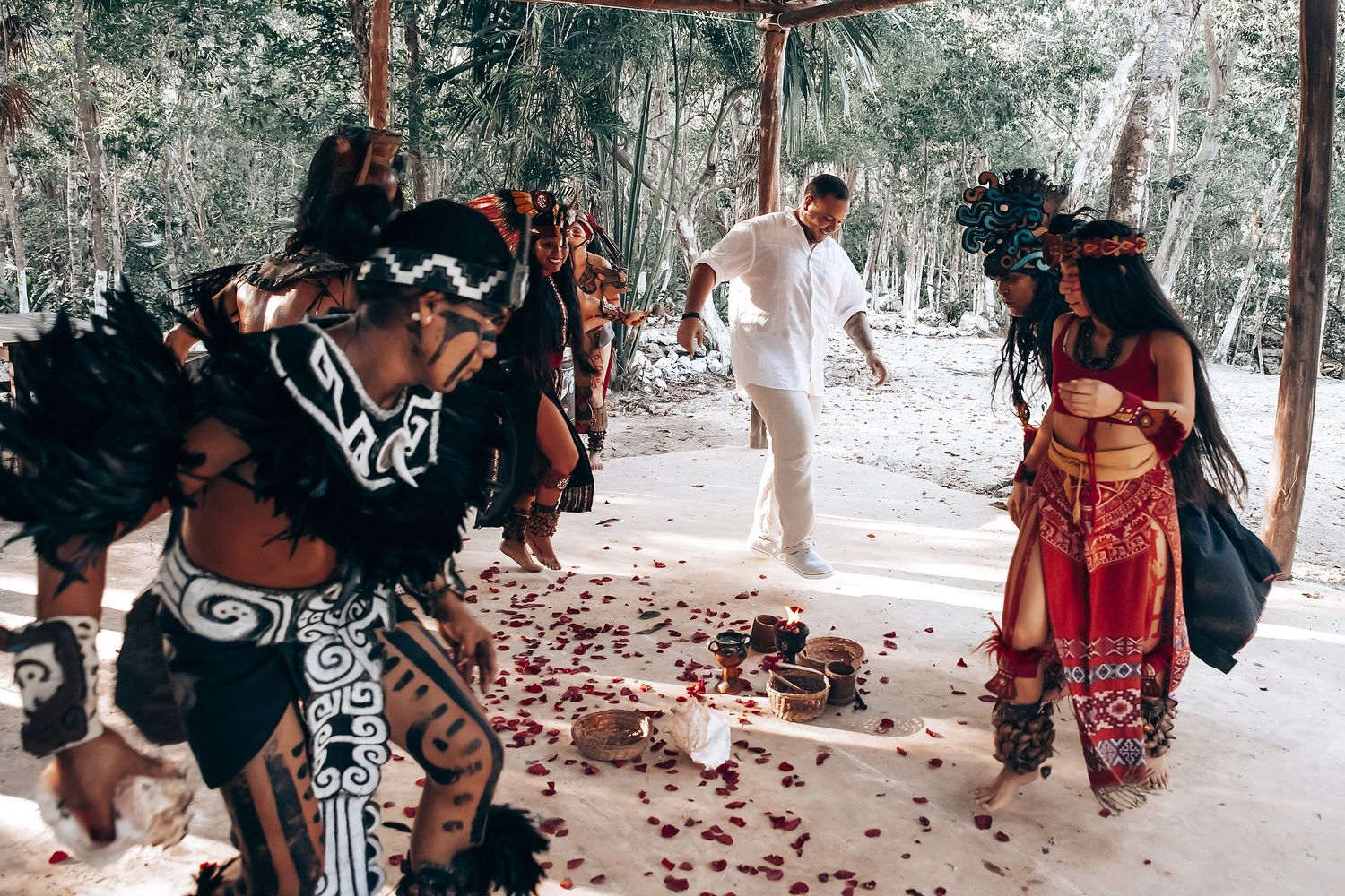 Traditional ceremony participants gathered in wooded area with rose petals scattered on ground.