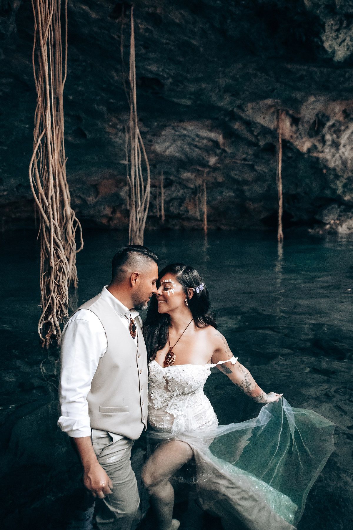 A couple shares an intimate moment in a water-filled cave with hanging vines and moody lighting.