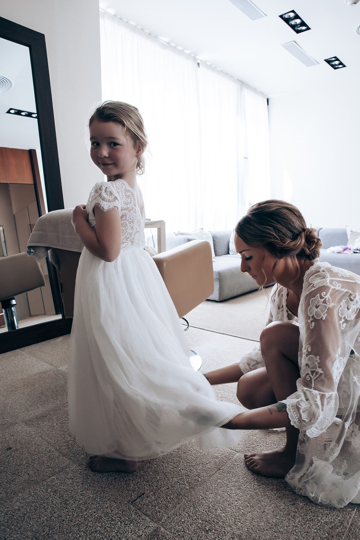A touching moment between a bride and flower girl in white lace dresses on a carpeted floor.