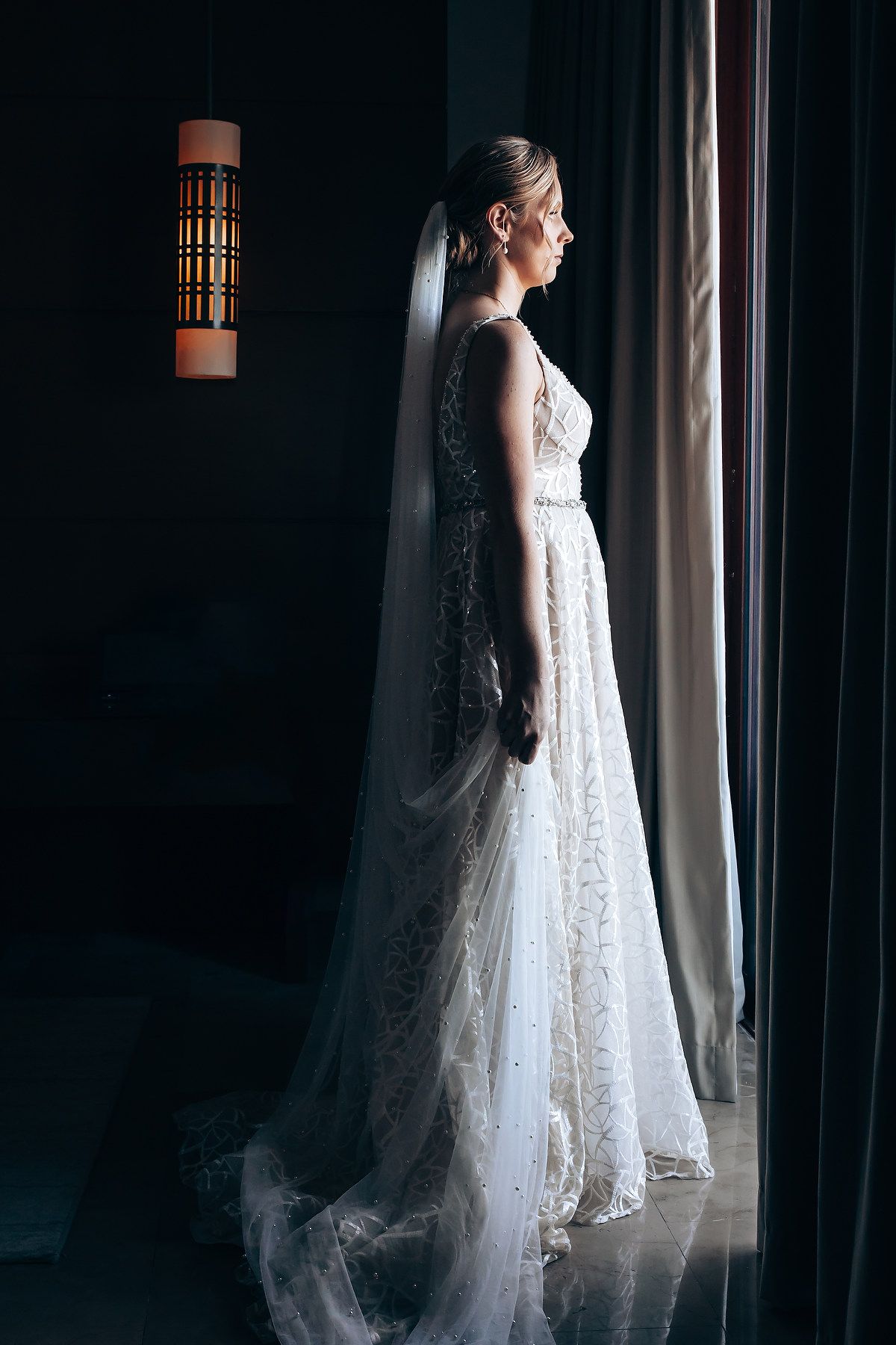 A bride standing in a dimly lit room, wearing a long, white lace wedding dress.