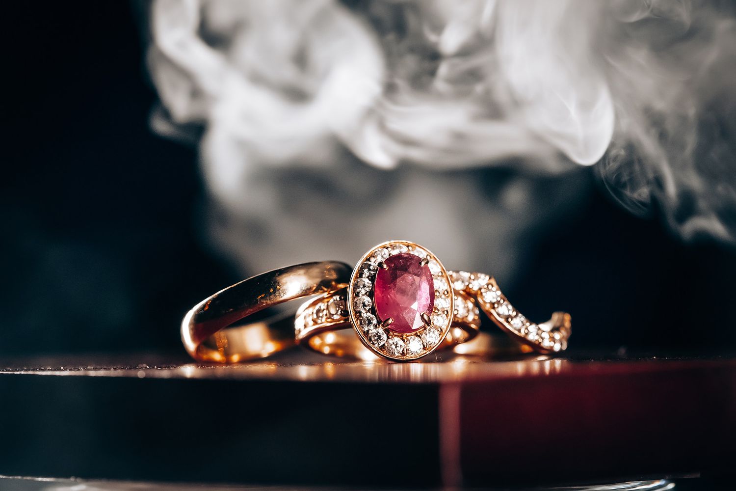A close-up view of a gold ring with a red gemstone and small diamonds, surrounded by smoke.